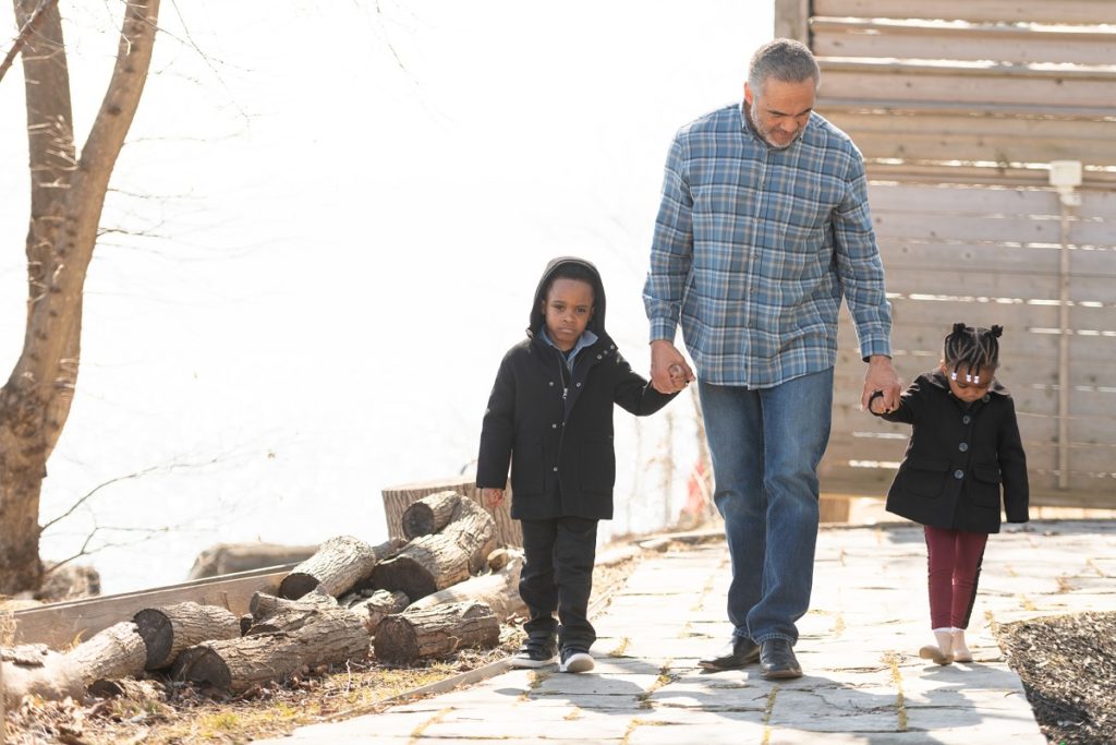 A man is playing with his grandchildren outside. The toddler girl and boy hold his hand as they all walk down a beautiful path together.
