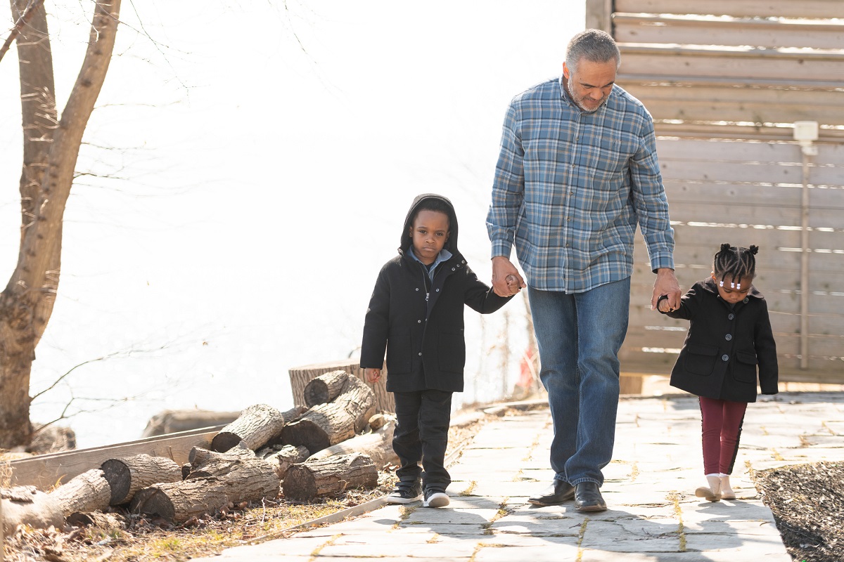 A man is playing with his grandchildren outside. The toddler girl and boy hold his hand as they all walk down a beautiful path together.