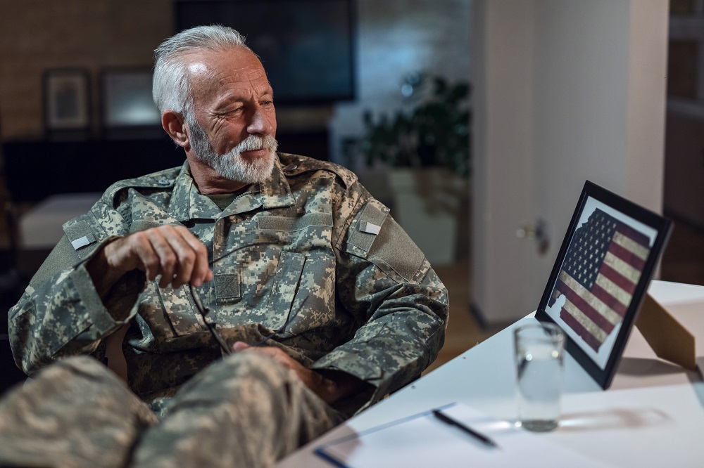 Baker Law Group - Veterans benefits Smiling mature army soldier feeling proud and admiring the American flag in picture frame while relaxing in the office.
