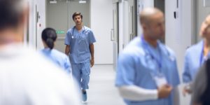 Doctor wearing blue scrubs and stethascope walking through hospital hallway