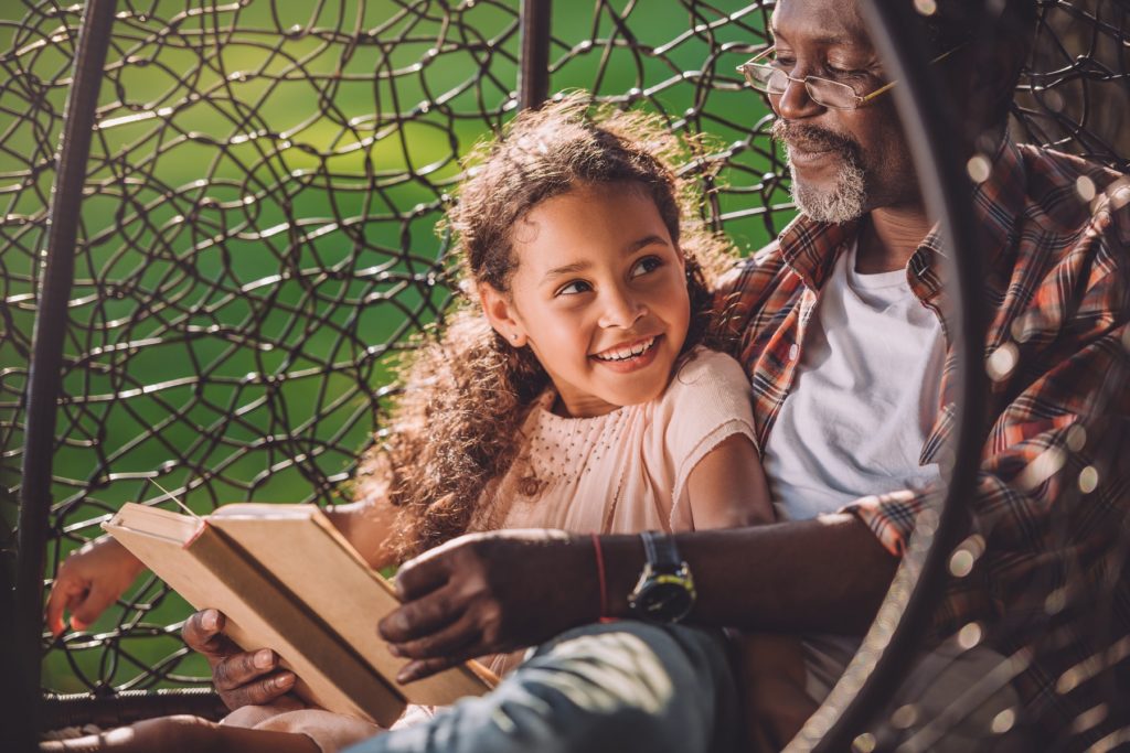 smiling granddaughter reading book while sitting in swinging hanging chair with grandfather