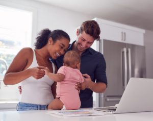 Family With Baby Daughter In Kitchen Using Laptop On Counter