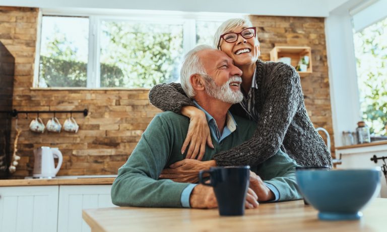 In their kitchen, a happy senior man is sitting and his smiling wife has her arms wrapped around him