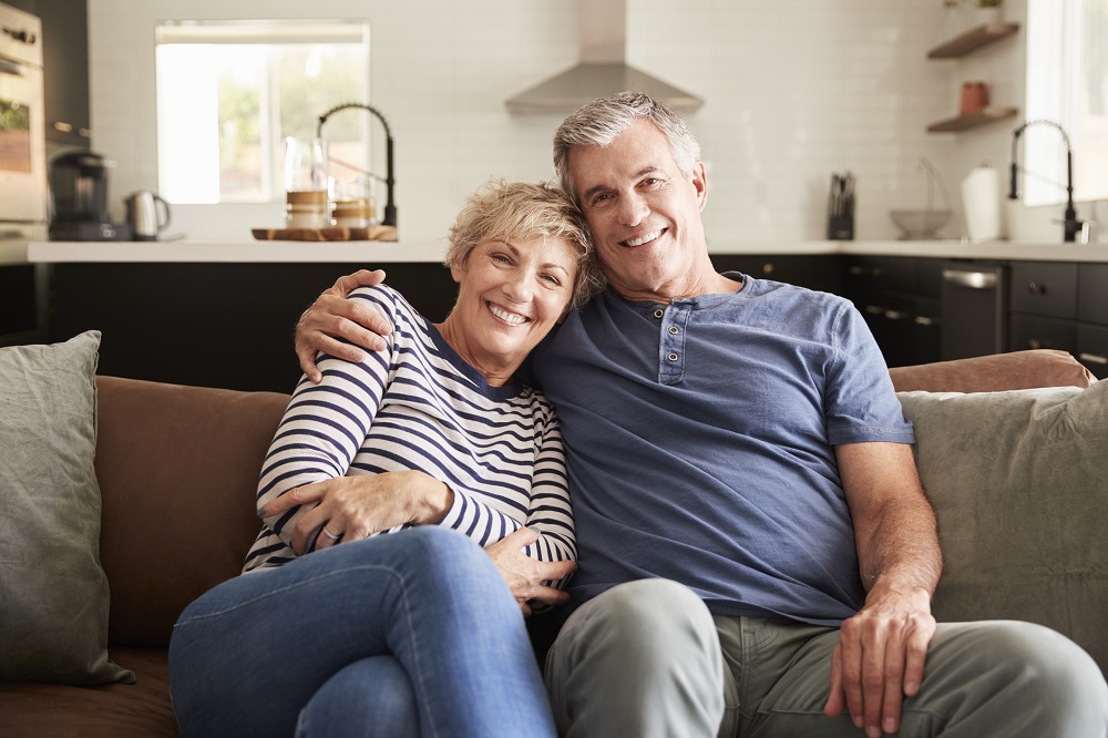 Mature couple sitting on couch with kitchen behind them, smiling into the camera