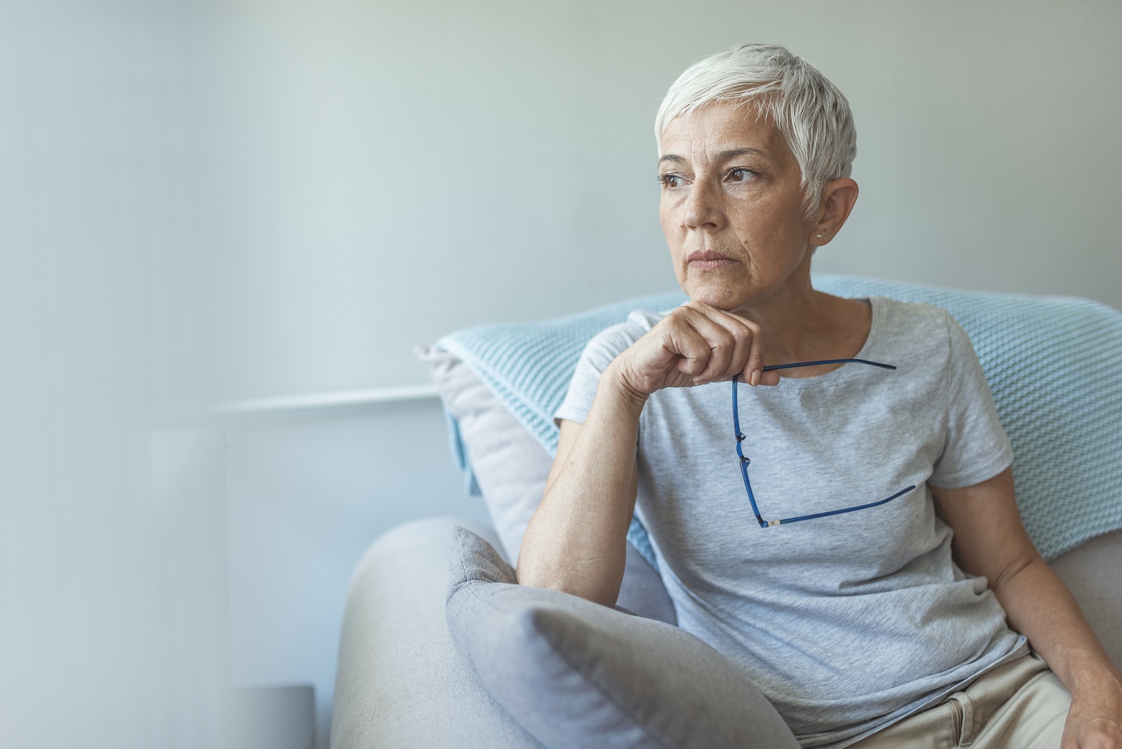 Mature woman sitting on couch looking thoughful, holding her glasses in her hand