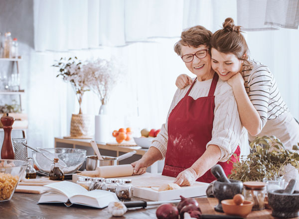 Grandmother in kitchen wearing red apron and rolling out a pie crust, smiling, while being hugged by her daughter who also has apron and smile