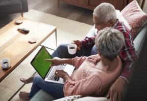 Senior Couple Sitting On Sofa At Home Using Laptop to eo online research