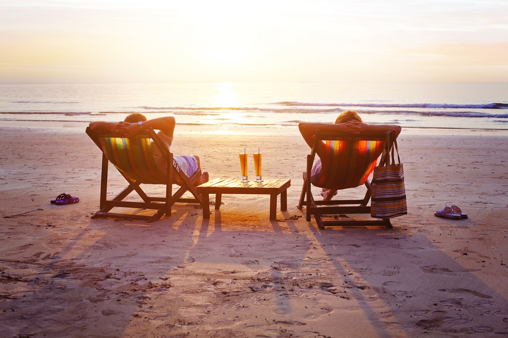 Couple fin lounge chairs facing the ocean and the sunset, with hands behind their head relaxing and having a glass of champagne