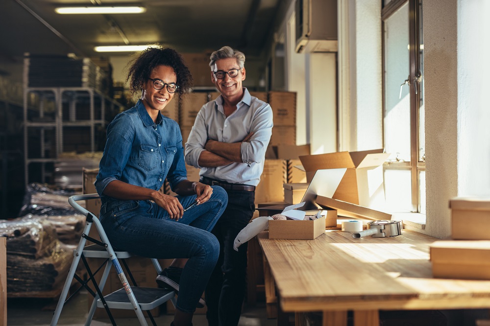 Smiling online store business partners. Woman sitting on a stool and man leaning against a counter looking at the camera