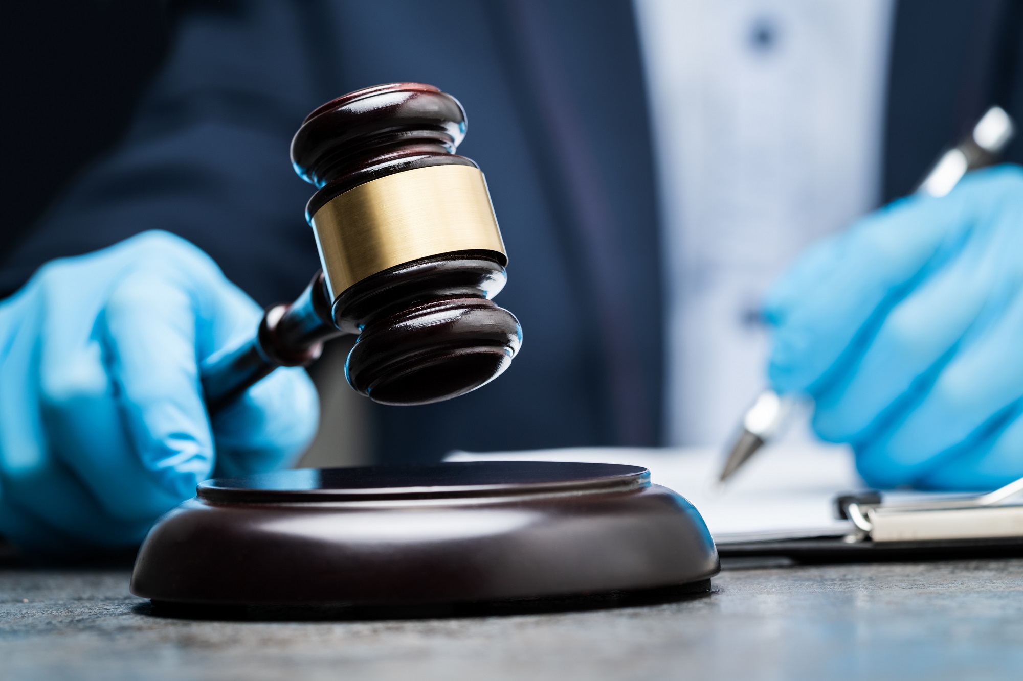 Close shot of a Judge's hands wearing protective blue gloves. He holds a pen and gavel.