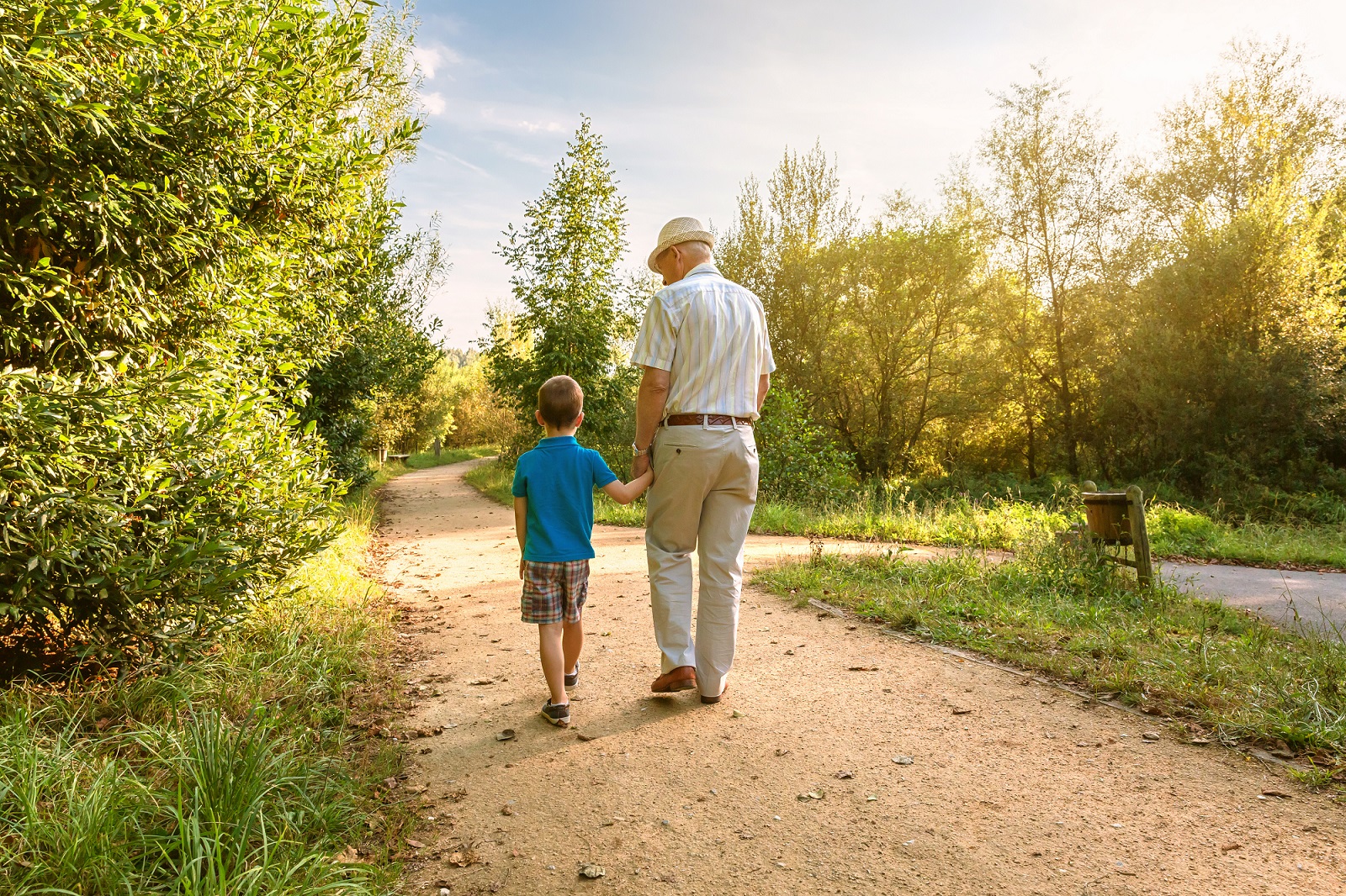 The back view of grandfather with hat and grandchild walking on a nature path