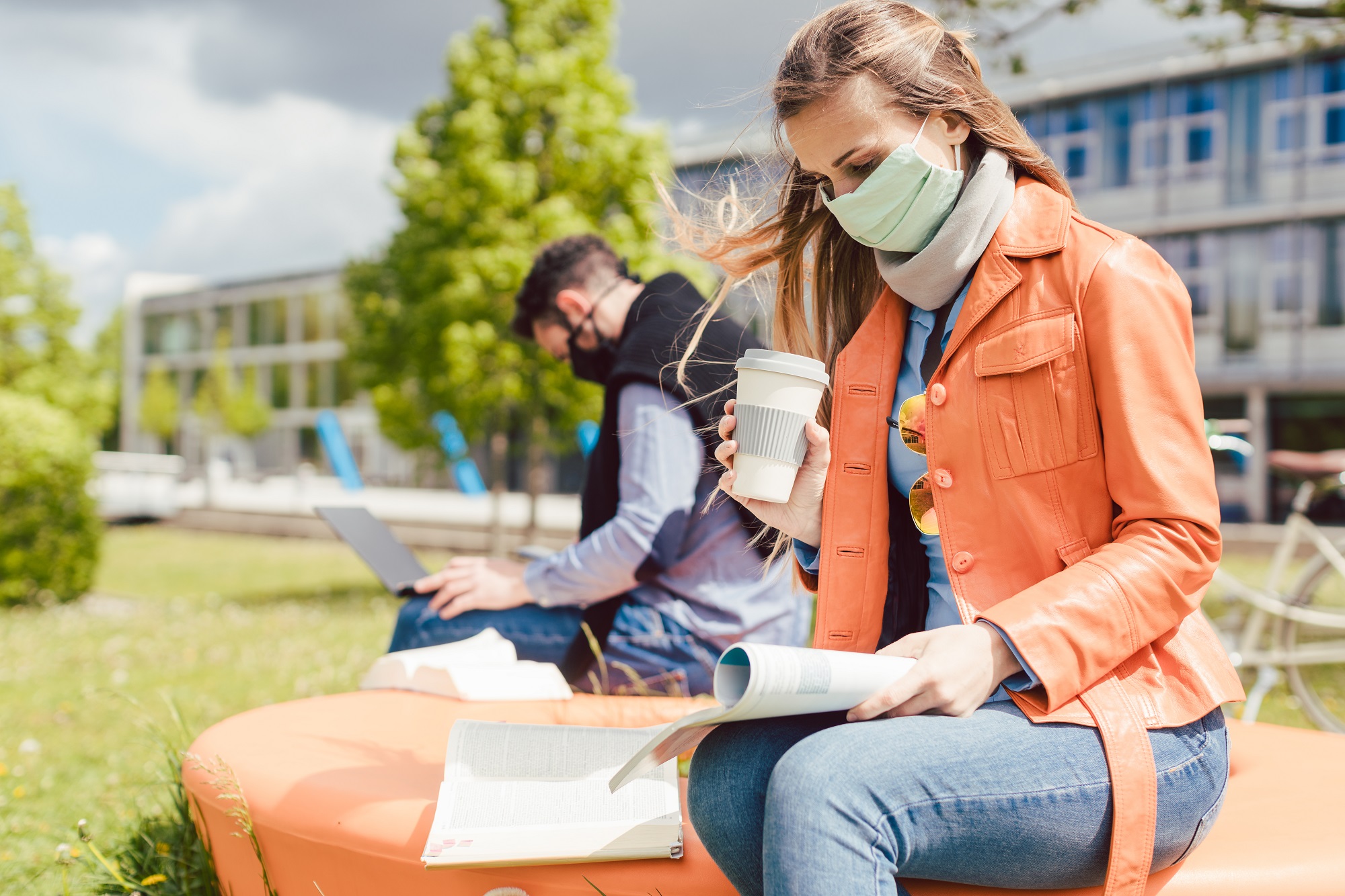 Man and woman student on college campus. Sitting on bench outside, wearing face masks and working on laptops