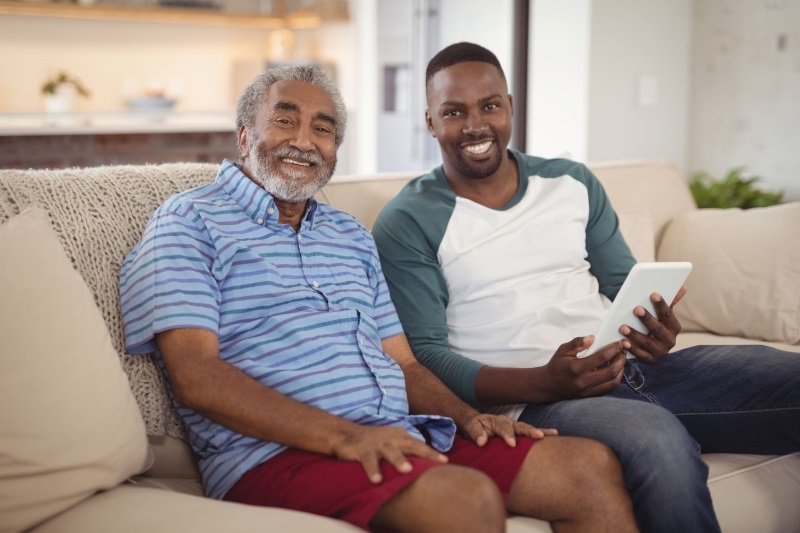Elderly man sitting on a couch with his son, both smiling and looking to the camera
