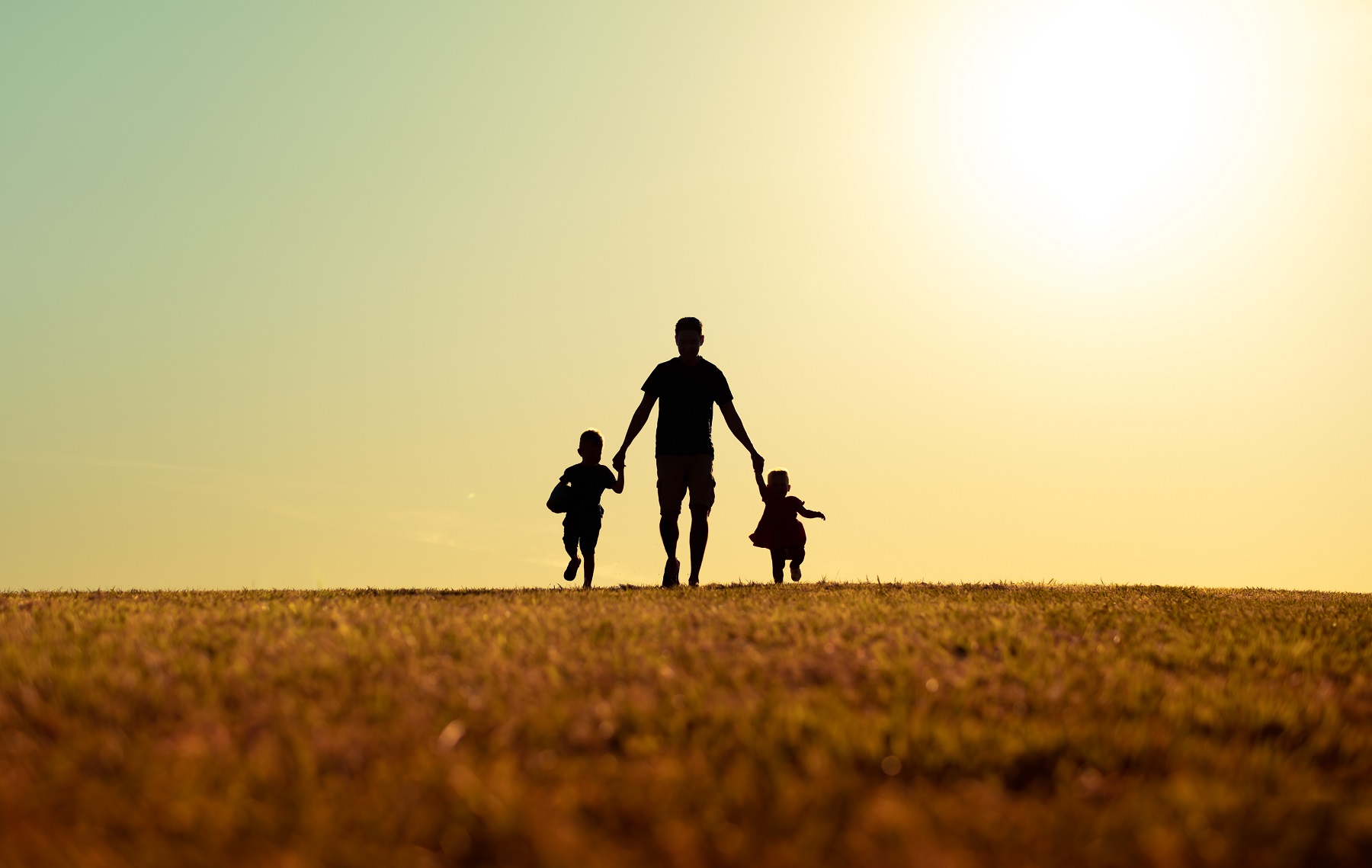 A silouette of a father walking away from the camera and holding hands with son and daughter in park at sunset.
